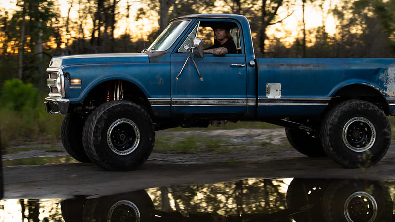 Chevrolet C20 lifted classic pickup driving off-road through a muddy trail at sunset.