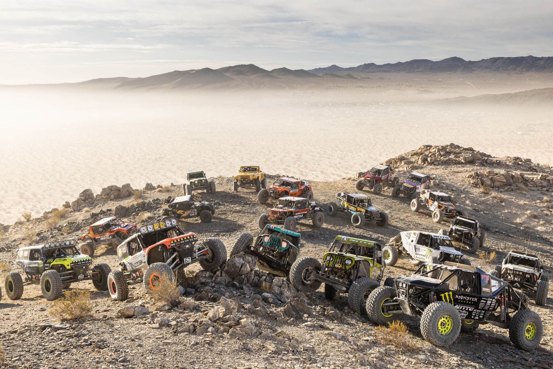 Group of rock crawler off-road vehicles lined up on a rocky hill during King of the Hammers race,
				with a desert landscape and mountains in the background.